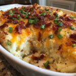 A scoop has been taken out of a baked mashed potato casserole topped with melted cheddar cheese, bacon bits, and green onions.