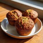 Three freshly baked, golden brown zucchini muffins resting on a light ceramic plate near a window.
