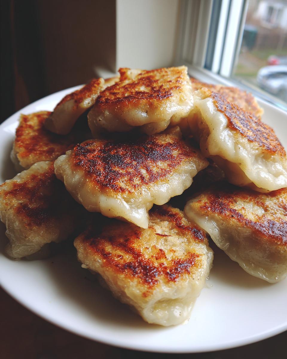 A close-up of several golden-brown, pan-fried Cabbage Dumplings piled high on a white plate near a window.