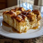 A close-up of a moist slice of Paula Deen Overnight French Toast Casserole drizzled with syrup on a white plate.