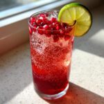 Close-up of a tall glass filled with a fizzy, red Pomegranate Spritz, topped with fresh pomegranate seeds and a lime wheel.