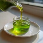 A stream of bright green apple syrup is being poured from a glass pitcher into a small jar sitting on a white saucer.