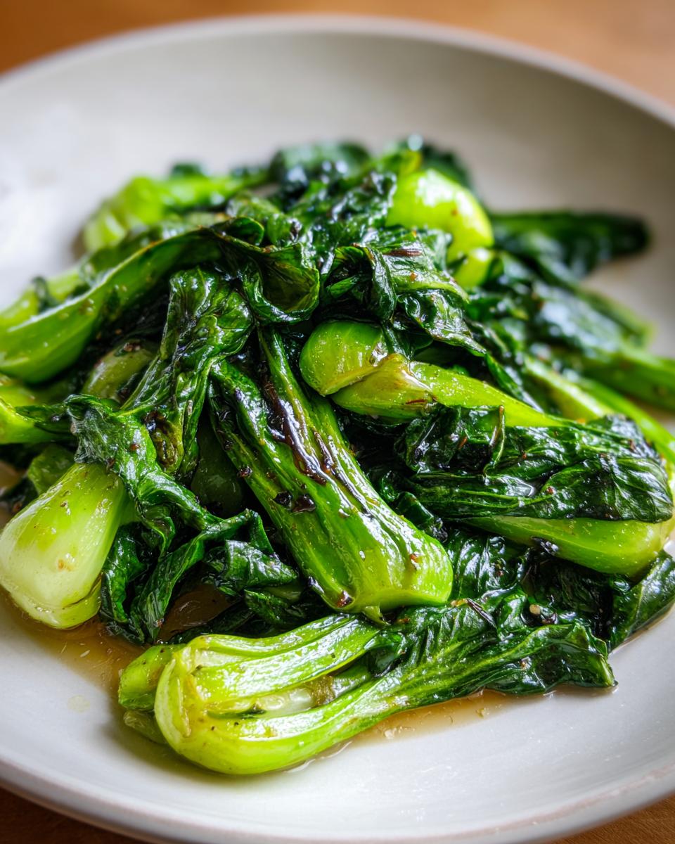 A close-up of vibrant green, glossy Sautéed Bok Choy served on a light-colored plate with sauce.