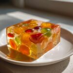A shimmering, amber-colored cube of Agar Agar Fruit Jelly filled with colorful pieces of preserved fruit, sitting on a white plate.