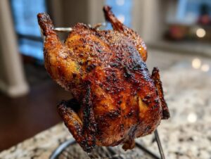 A whole, beautifully browned and glazed smoked beer can chicken standing upright on a metal rack.