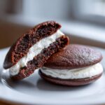 Two rich chocolate whoopie pies on a white plate, one cut in half showing the soft cake texture and thick white cream filling.