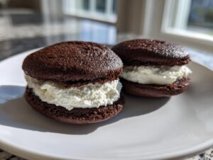 Two homemade chocolate whoopie pies filled generously with fluffy white cream frosting, sitting on a white plate.