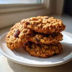 A stack of three golden brown oatmeal raisin cookies resting on a white plate, highlighted by bright sunlight.