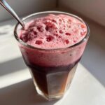 Close-up of a glass with a dark liquid topped with thick, bright pink Strawberry Cold Foam and a spoon.
