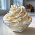 A generous swirl of fluffy, pale yellow vanilla buttercream frosting piled high in a clear glass bowl.