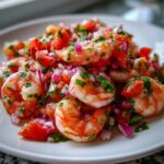 Close-up of bright, juicy shrimp ceviche mixed with diced red onion, tomato, and cilantro on a white plate.