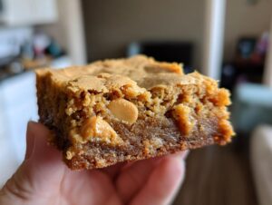 A hand holds up a thick, fudgy square of butterscotch brownies showing melted butterscotch chips inside.