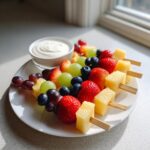 Three colorful fruit skewers featuring strawberries, pineapple, grapes, and blueberries, served with a side of yogurt dip.