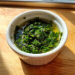 Close-up of vibrant green, freshly chopped mint sauce resting in a small white ramekin on a wooden surface.