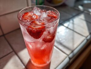 Close-up of a tall, sweating glass filled with bright red Strawberry Lemonade, ice cubes, and fresh strawberry slices.