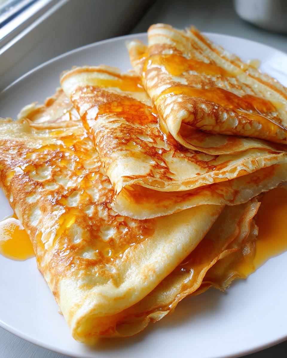 Close-up of folded Crepes Suzette stacked on a white plate, glistening with orange syrup.