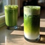 Two tall glasses of iced Matcha latte, one layered with milk and the other fully mixed, sitting on a sunlit counter.