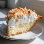 A tall slice of creamy Coconut cream pie topped with whipped cream and toasted coconut flakes.
