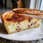 A thick slice of creamy Quiche Lorraine, showing bacon pieces in the custard filling, with the whole quiche blurred in the background.