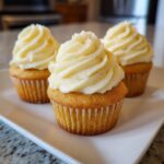 Three perfectly baked lemon cupcakes topped with thick swirls of pale yellow frosting, resting on a white plate.